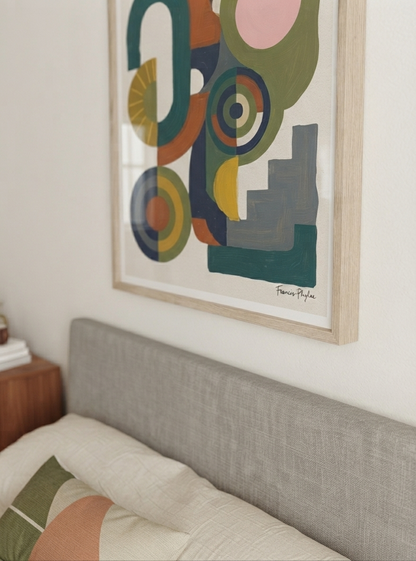 A framed Bauhaus Abstract Geometric Wall Poster with retro shapes art print for living room by Francis Phylae from Phylae Posters, with geometric shapes in muted green, teal, yellow, orange, pink, and gray, hanging on a white wall above a gray upholstered bed headboard.