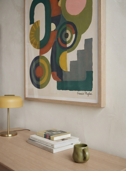 A framed Bauhaus Abstract Geometric Wall Poster inspired by architectural shapes poster design from Francis Phylae at Phylae Posters, with mid-century geometric shapes in green, orange, yellow, pink, and gray, hanging on a textured off-white wall above a light wood console table with a yellow lamp, books, and a green ceramic mug.