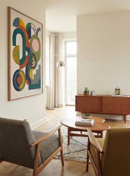 A modern living room interior with a large framed Bauhaus Abstract Geometric Wall Poster inspired by Scandinavian geometric wall decor unframed from original artwork by Francis Phylae at Phylae Posters on the wall, a mid-century wooden credenza, two upholstered armchairs, a round coffee table, and a window with sheer curtains.