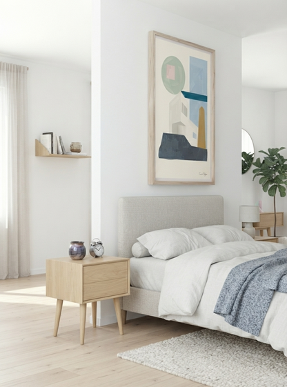 A modern bedroom scene featuring a light wood nightstand, a bed with white bedding and a blue throw blanket, and a large framed Mid-Century Modern Abstract House Print featuring minimalist Bauhaus-inspired architecture in calming sage green and blue tones by Francis Phylae on the wall above the headboard.