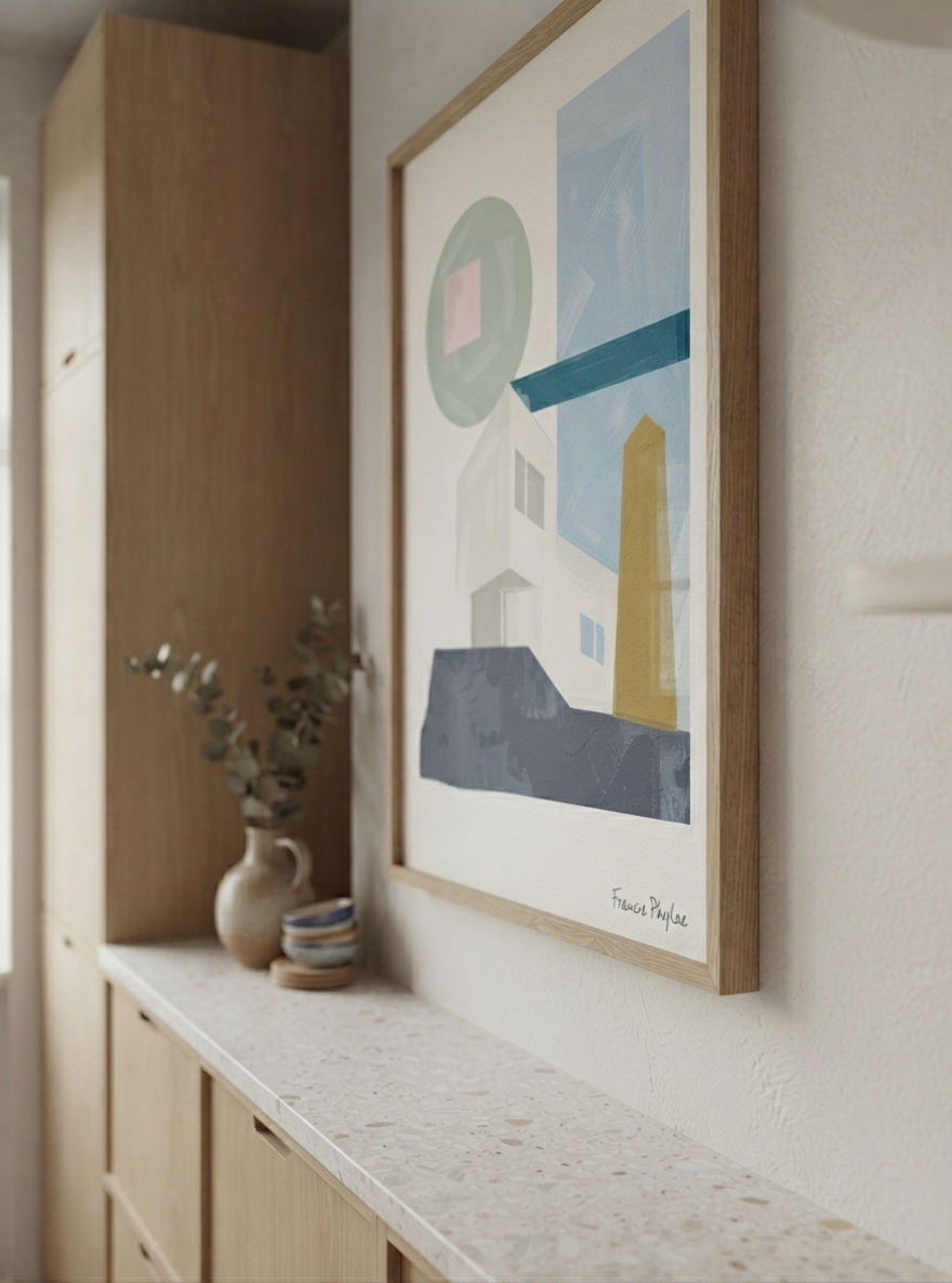 A framed Affordable Mid-Century Home Decor Art with geometric architectural composition, created from original acrylic artwork by Francis Phylae,  hangs on a white wall above a light wood console table. The print features minimalist shapes in blue, gray, white, yellow, and green on a white background, enclosed in a light natural wood frame.