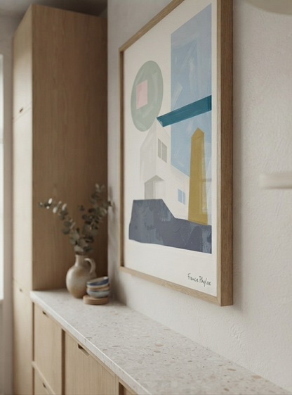 A framed Affordable Mid-Century Home Decor Art with geometric architectural composition, created from original acrylic artwork by Francis Phylae,  hangs on a white wall above a light wood console table. The print features minimalist shapes in blue, gray, white, yellow, and green on a white background, enclosed in a light natural wood frame.