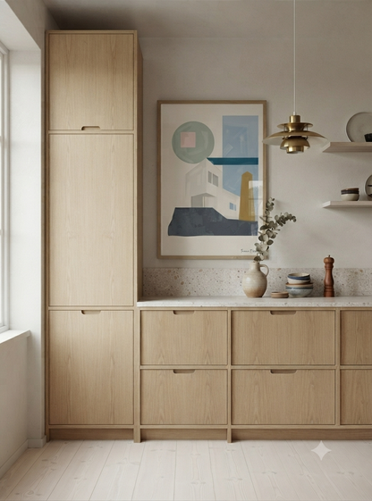 A modern kitchen interior featuring light oak wood cabinetry, including a tall pantry cabinet and lower drawers with matching fronts, a white countertop, a framed Modern Architectural Poster for Living Room featuring geometric architectural forms inspired by Bauhaus design and Mid-Century Modern aesthetics on the wall, a hanging brass pendant light, and a vase with dried stems.