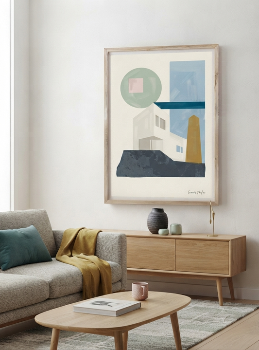 A framed abstract Mid-Century Modern Architectural Art Print inspired by Bauhaus architecture, showcasing a minimalist abstract house print from original acrylic artwork by Francis Phylae, hangs on a white wall above a light wood sideboard in a modern living room setting. The print features white, blue, green, and yellow shapes, and the room includes a gray sofa with a yellow throw blanket, a wooden coffee table, and a small vase on the sideboard.