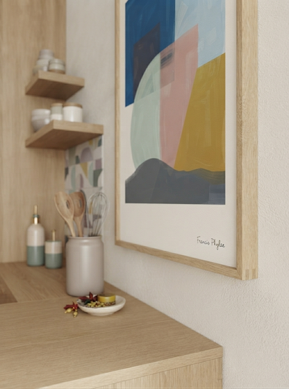 A framed Mid-Century Modern Geometric Landscape Art Print featuring grey and blush abstract painting tones by Francis Phylae, available from Phylae Posters with geometric shapes in blue, pink, yellow, and gray, hanging on a light wall above a wooden desk with kitchen accessories.