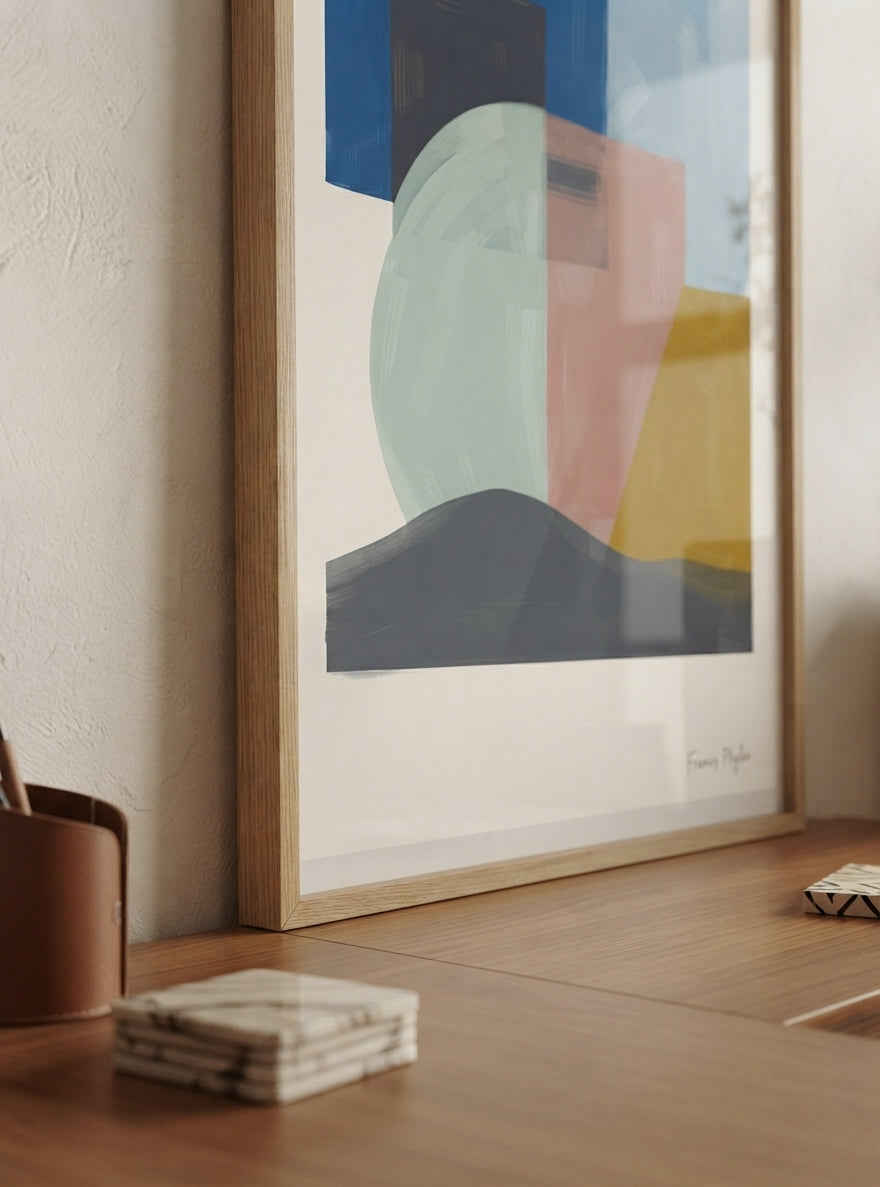 A framed Mid-Century Modern Geometric Landscape Art Print featuring navy blue and dusty rose art print colours from an original painting by Francis Phylae for Phylae Posters with geometric shapes in blue, mint green, pink, yellow, and dark gray, leaning on a wooden table next to a textured wall.