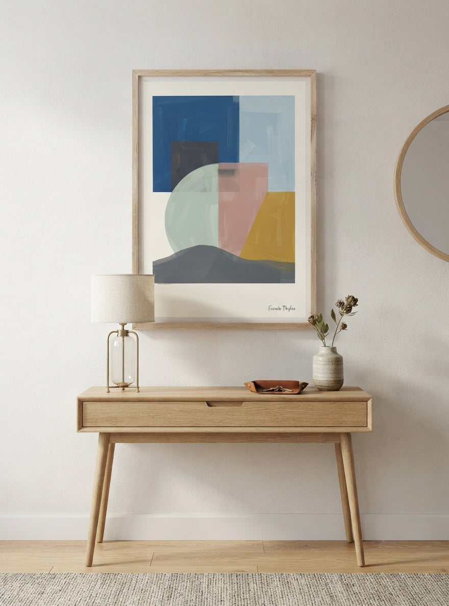 A framed Mid-Century Modern Geometric Landscape Art Print styled as a Scandinavian Art Print with modern abstract shapes by Francis Phylae for Phylae Posters hangs above a light wood console table in a bright, modern room. The artwork features blocks and curves in blue, pink, yellow, gray, and white, with a light natural wood frame.
