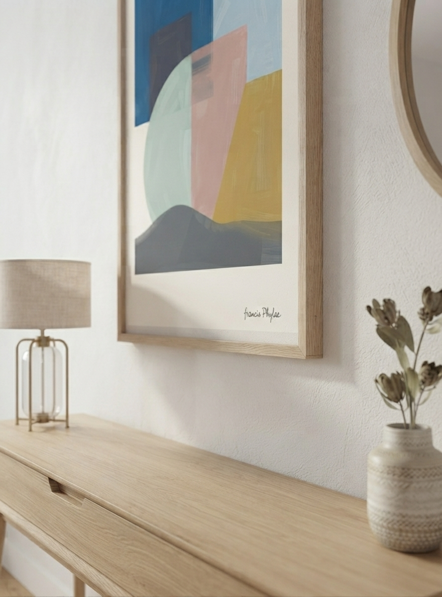 A framed Mid-Century Modern Geometric Landscape Art Print styled as a Scandinavian Art Print with modern abstract shapes by Francis Phylae for Phylae Posters featuring geometric shapes in blue, pink, yellow, and gray, hanging on a white wall above a light wood console table with a lamp and a vase.