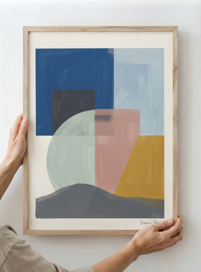 A framed Mid-Century Modern Geometric Landscape Art Print inspired by contemporary geometric landscape canvas art from original acrylic artwork by Francis Phylae for Phylae Posters held by a person's hands against a plain white wall. The print features overlapping blocks and shapes in blue, pink, yellow, gray, and pale green, set in a light natural wood frame.